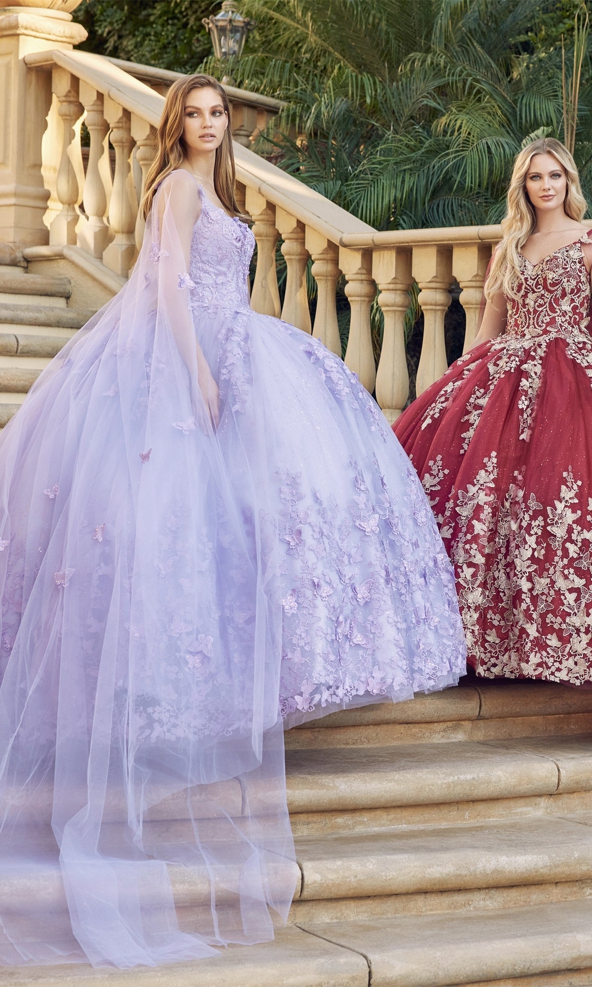 Two young women stand on outdoor stairs; one wears a lavender floral gown, the other showcases the Juliet 1442 Quinceañera Dress with Sheer Cape—a stunning red embroidered dress with gold accents.